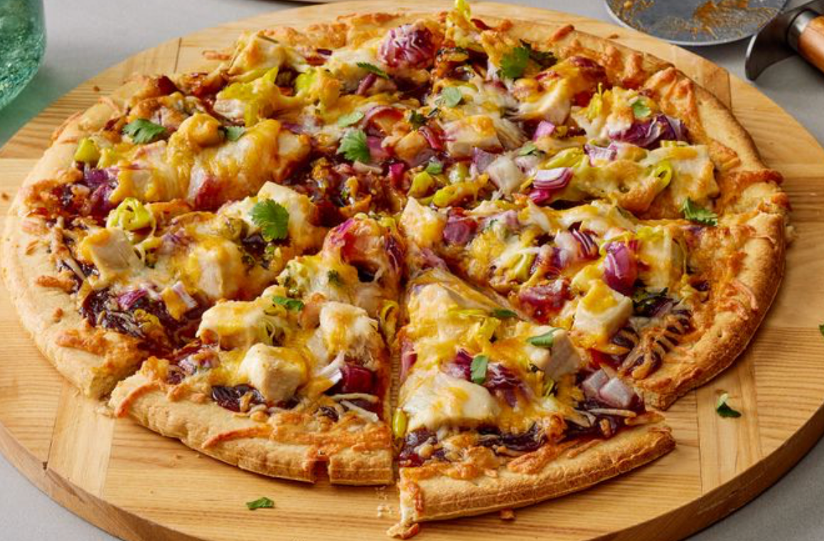 A large cut pizza with melted cheese, chicken, red onion, green peppers, and herbs, served on a round wooden board.